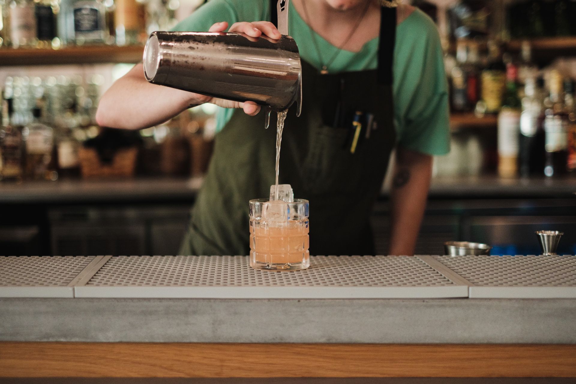 A bartender pours a cocktail from a shaker into a glass at a bar counter.